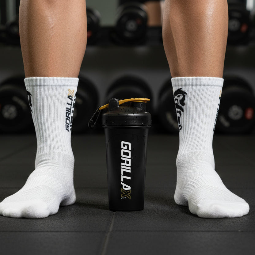 Person wearing white socks with 'Gorilla' branding, standing on a gym floor with a black shaker bottle.